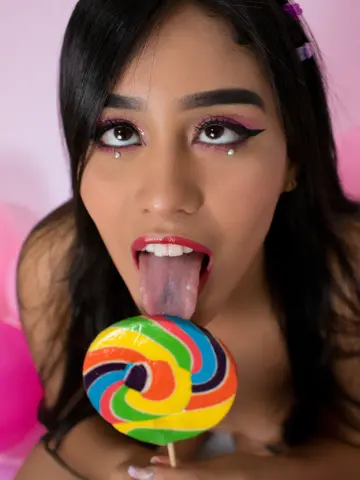 Young woman with dark hair and winged eyeliner playfully eating a colorful swirl lollipop against a pink background.