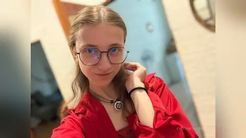 Young woman with glasses and a red shirt smiling in a bright indoor setting.