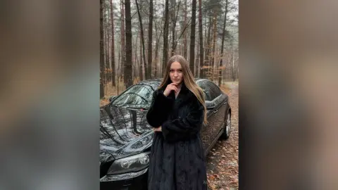 AuroraCruise poses in a long black coat standing next to a black car in a forest setting.
