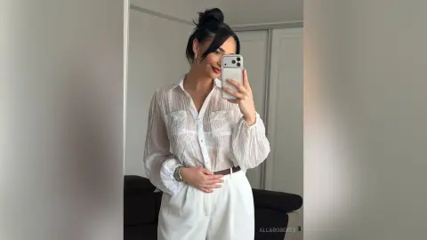 Young woman in a white sheer blouse and trousers taking a mirror selfie in a bright modern room.