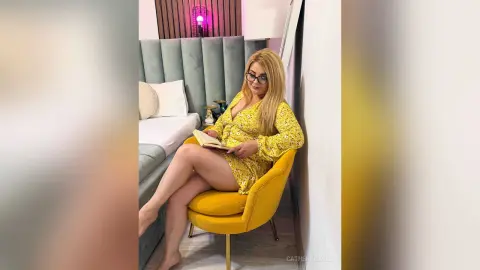 Blonde woman in a yellow floral dress reading a book on a yellow velvet chair in a modern living room.