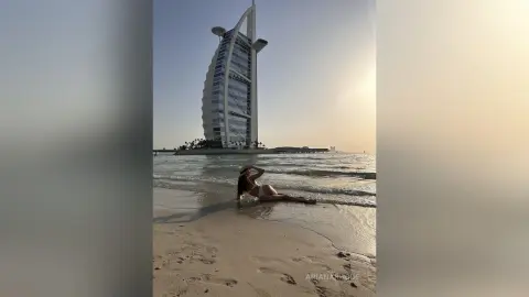 Woman in white bikini relaxing on a sandy beach with the Burj Al Arab hotel in the background at sunset.