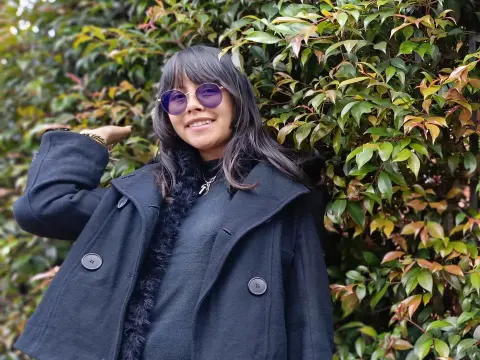 Smiling woman in a black wool coat and purple sunglasses standing in front of lush green garden foliage.