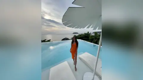 MilanaVolkova stands in an orange dress next to an infinity pool overlooking the ocean under a white umbrella.