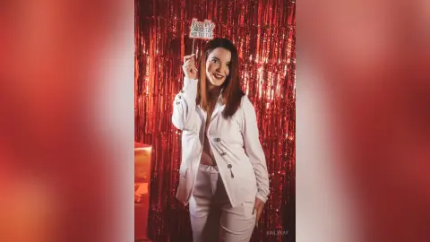 Smiling woman in white outfit holding a Kiss Me Under the Mistletoe sign against a festive red sequin backdrop.