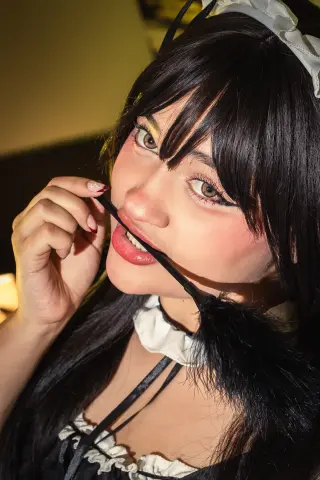 Smiling young woman in a black and white maid cosplay outfit with dark hair and dramatic eye makeup.