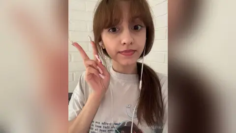 Young woman with brown hair wearing a white graphic t-shirt and wired earbuds making a peace sign indoors.