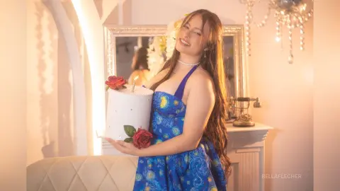 Smiling woman in a blue floral dress holding a white birthday cake in a warm, sunlit room.