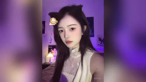 HedyIvy poses with long dark hair and cat ears, wearing a white lace sleeveless top against a purple lit background.
