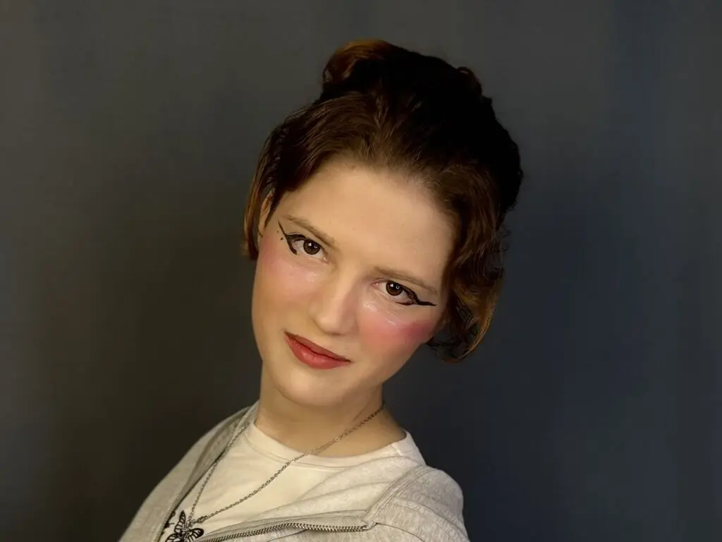 IrisKemp poses with her hair in a bun and heavy winged eyeliner, wearing a white shirt and silver necklace against a dark grey background.