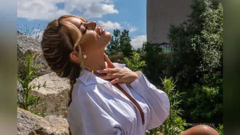 Blonde woman in a white button-down shirt relaxing outdoors against a sunny sky and green trees.