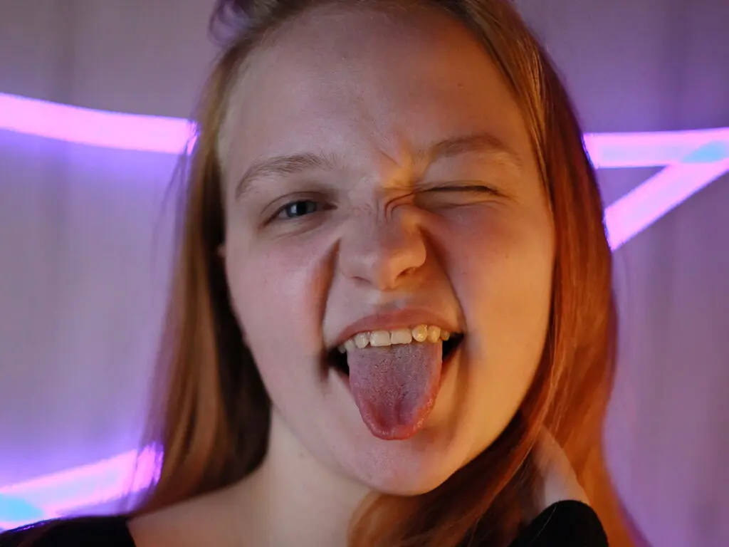 Playful redhead girl making a funny face and sticking out her tongue in a room with purple neon lighting.