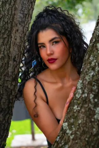 Young woman with curly dark hair in a black strap dress posing between tree trunks in a sunny outdoor setting.
