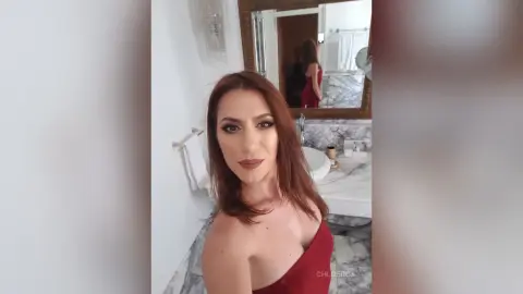 Redheaded woman in a red dress posing in a bright marble bathroom with a confident expression.