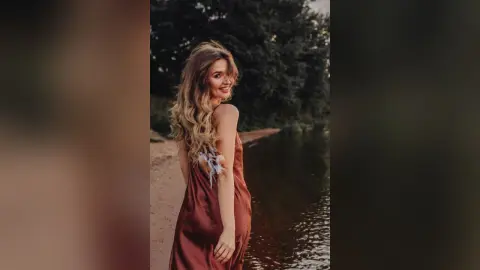 Smiling blonde woman in a brown satin dress standing by a serene lake at sunset.
