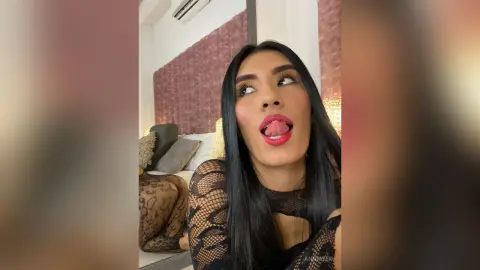 AnnMyer licks her lips while wearing a black lace long-sleeve top, looking off-camera with long black hair in a room with a textured wall and sofa.