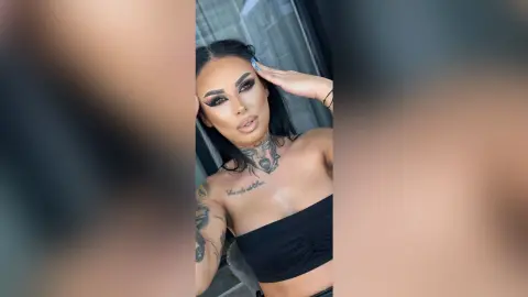 Woman with dramatic winged eyeliner and tattoos wearing a black tube top in a moody indoor setting.