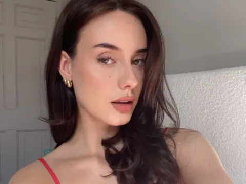 Young woman with long brown hair and gold hoop earrings wearing a red camisole in a bright indoor setting.