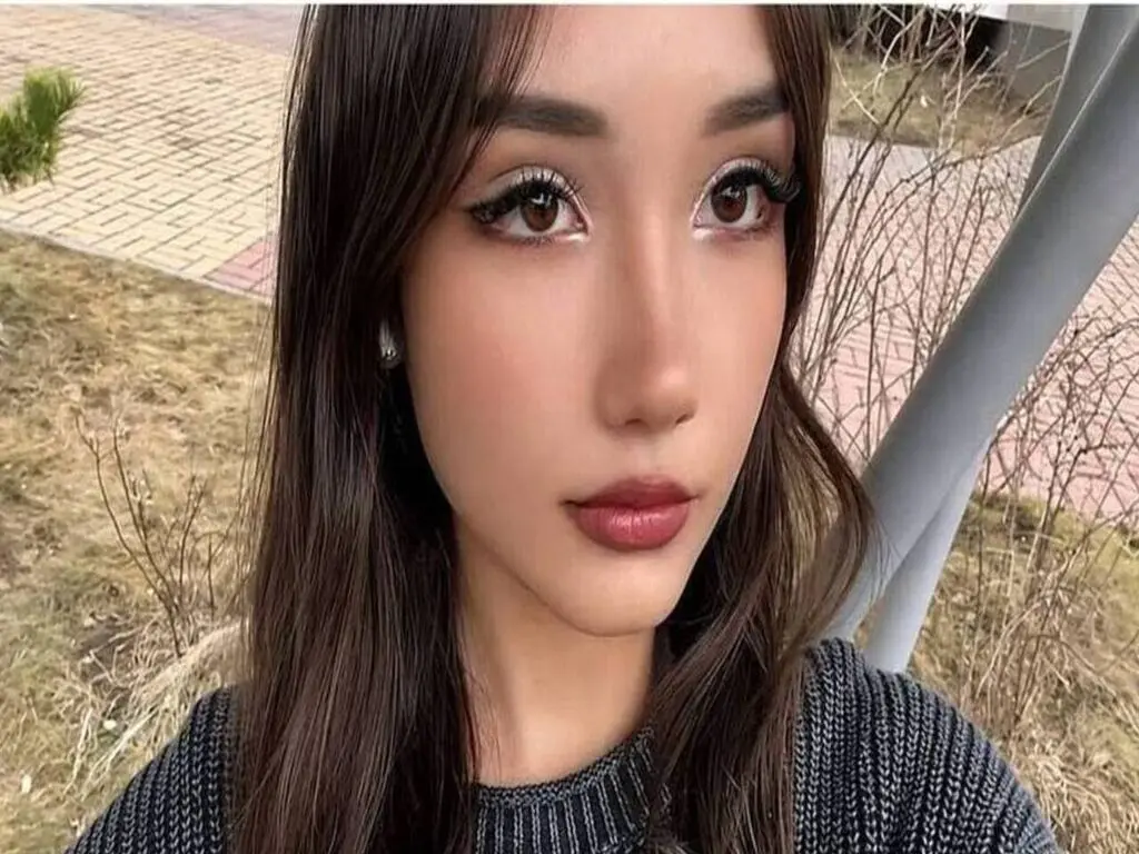 Close-up portrait of a young woman with long dark hair and glamorous eye makeup in an outdoor setting.