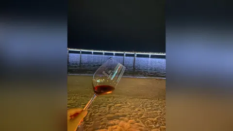 KimFu — KimFu holds a glass of red wine on a sandy beach at night with a lit bridge in the background.