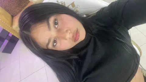 Young woman with long dark hair wearing a black turtleneck sweater in a bright indoor setting.