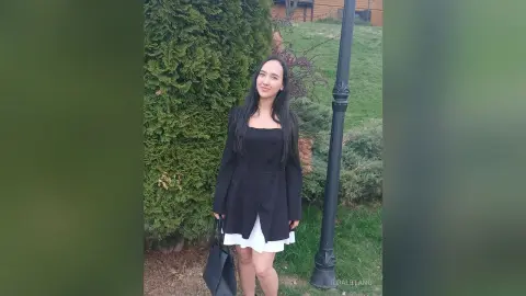 Smiling young woman in a black long-sleeve dress standing in a lush green garden by a lamp post.