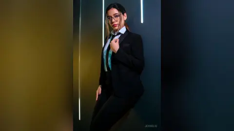 JessieLiu wears a black suit and tie with a light blue shirt, posing against a dark wall with vertical light strips.