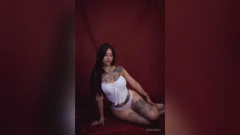 OrianaWill poses in a white lace corset and matching undies, showing off her tattoos against a dark red background.