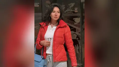 MnemoZeina wears a red puffer jacket over a white shirt and grey sweatpants, holding a pair of glasses and a blue bag.