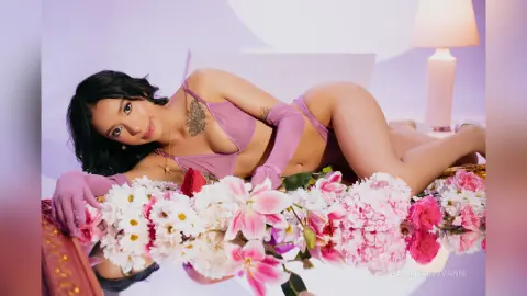 Smiling woman in lavender lingerie and gloves lying on a bed of pink and white flowers in a soft dreamy setting.