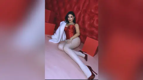 AnnaKarev sits on a pink floor in a red corset and white thigh-high stockings against a red tufted wall.