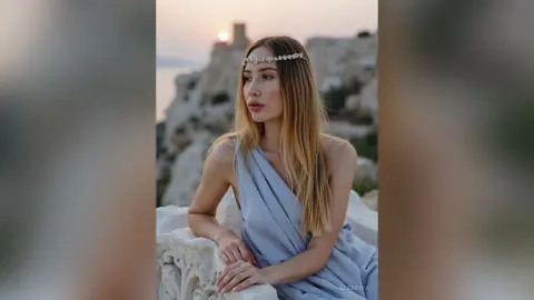 Elegant woman in a light blue Grecian gown wearing a silver headband at a scenic coastal ruin during sunset.