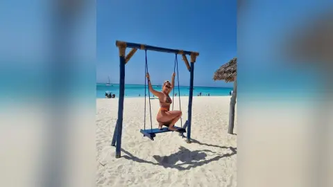 Blonde woman in a brown bikini relaxing on a wooden swing at a sunny tropical beach with turquoise water.