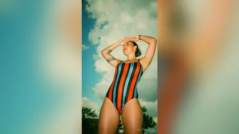 Woman in a colorful striped one-piece swimsuit posing under a bright blue sky with a carefree summer mood.