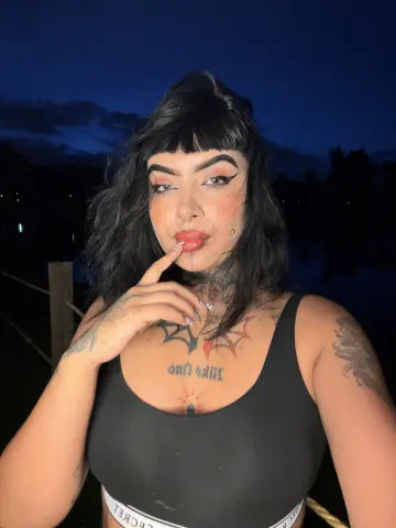 Woman with black hair and tattoos wearing a black tank top outdoors at dusk.