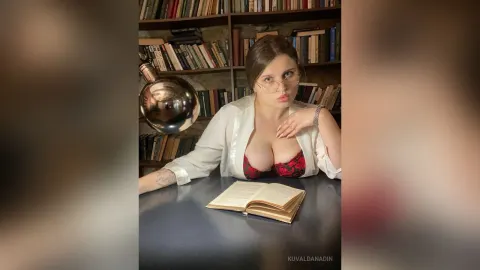 KuvaldaNadin — KuvaldaNadin wears an open white shirt and red lace bra while sitting at a desk with an open book in front of a bookshelf.