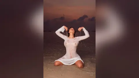 Elie poses wet in a tight white long-sleeve dress while kneeling on a beach at sunset, lifting her arms above her head.