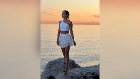 Young woman in a white summer outfit standing on coastal rocks during a serene golden hour sunset.