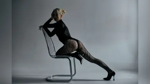 Blonde woman in a black bodysuit and patterned tights posing on a silver chair in a minimalist studio setting.