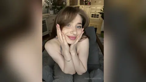Smiling young woman with brown hair in a grey tank top relaxing in a cozy indoor setting.