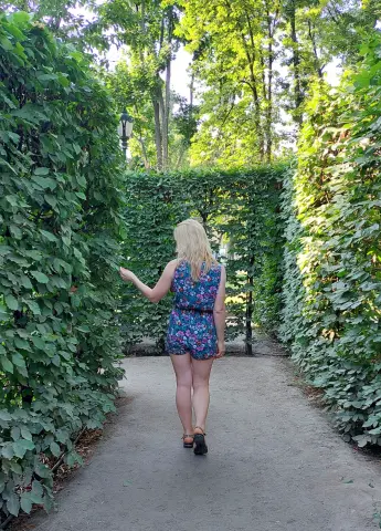 Blonde woman in a floral romper walking through a lush green garden path on a sunny day.