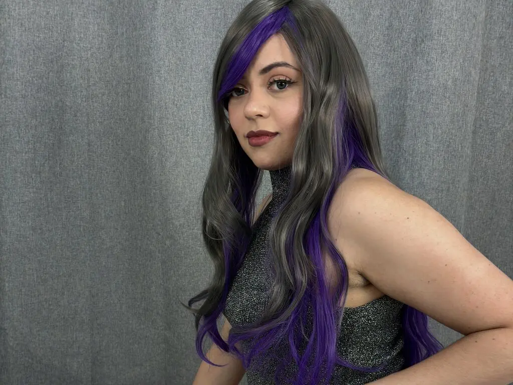 Woman with long wavy grey and purple hair wearing a sparkly silver halter top against a grey backdrop.