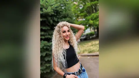 Smiling blonde woman in a black crop top and jeans posing outdoors in a lush green park.