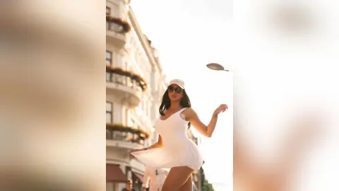 NicoleBella wears a white sundress and a white cap while standing outdoors against a bright sky.