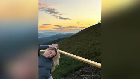 Blonde woman in a grey long-sleeve shirt relaxing on a balcony overlooking a scenic mountain sunset view.