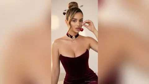Glamorous woman in a burgundy velvet corset holding a cherry against a soft neutral background.