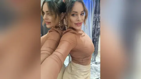 Glamorous woman in a tan long-sleeve top and hoop earrings posing for a mirror selfie in a boutique.
