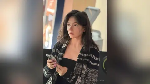 MnemoZeina has long dark hair and wears a black top with a black and white patterned cardigan while holding a phone.