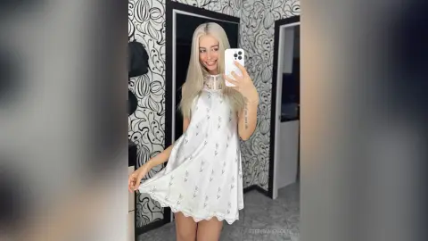 Smiling blonde woman in a white floral lace slip dress posing for a mirror selfie in a modern room.