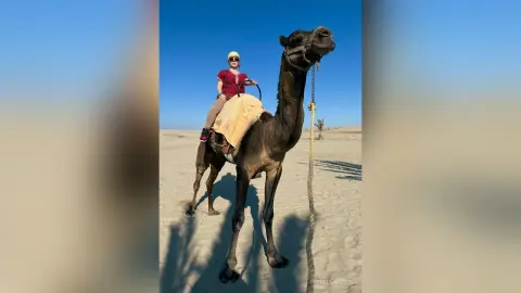 Smiling woman in casual summer attire riding a camel through a sunny desert landscape.